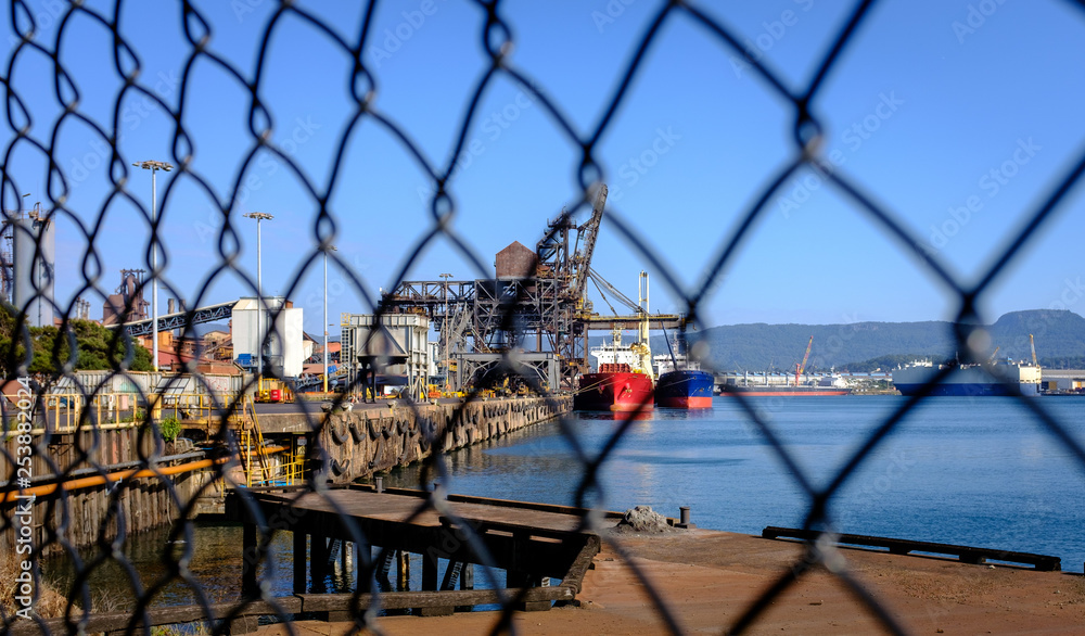 Ships loading coal and iron ore at industrial port taken through wire fence in foreground at an Australian Port, NSW. Trade war and trade tariffs concept.