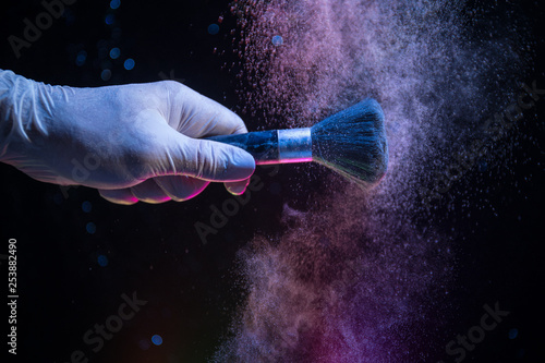 Makeup brush in hand with cosmetic powder on dark background with light and smoke. Powder splash on dark