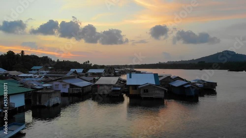 Ascending aerial view of small homes on stilts over calm water as the sun rises on Tawi Tawi, Philippines. Tropical landscape and peaceful morning scene. Mountain in the distance. photo