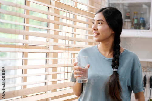 Youn beautiful asian woman drinking water while standing by window in kitchen background, peolpe and healthy lifestyles photo