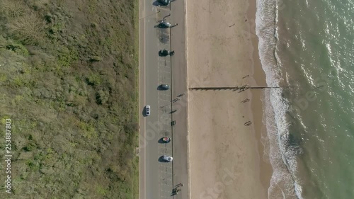 Topdown of a beachside road. One side is a big grass bank, on the other a golden sandy beach, and tropical murky ocean water with waves. Follows silver saloon car along a busy route. photo