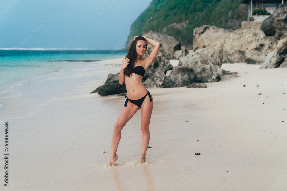Beautiful girl in a black swimsuit walks on the white sand beach near the ocean.