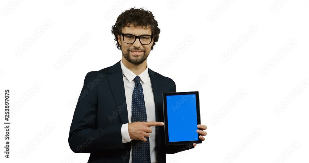 Caucasian good looking man in suit and tie posing with a black tablet computer in hands and pointing with his finger at it on the white wall background. Tracking motion Caucasian good looking man in suit and tie posing with a black tablet computer in hands and pointing with his finger at it on the white wall background. Tracking motion
