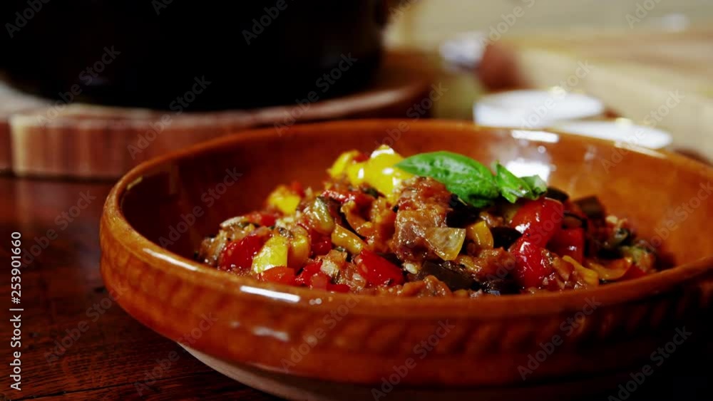 Video zooming in on cooked ratatouille recipe in a rustic ceramic dish sitting on a wooden country table. Video zooming in on cooked ratatouille recipe in a rustic ceramic dish sitting on a wooden country table.
