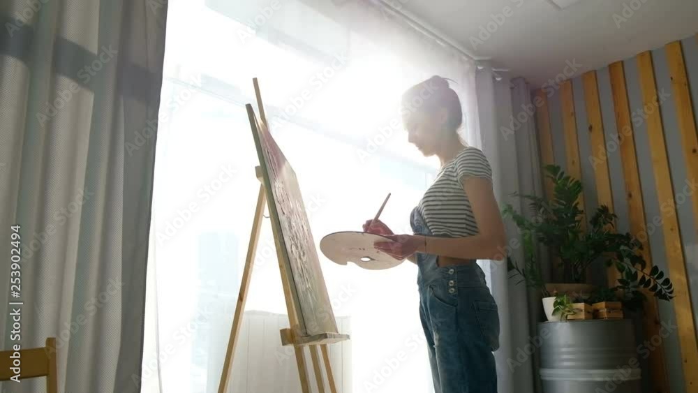 Young woman artist painting picture on easel in art studio. Talented girl creates a beautiful picture for the interior. art school, creativity and people concept Young woman artist painting picture on easel in art studio. Talented girl creates a beautiful picture for the interior. art school, creativity and people concept