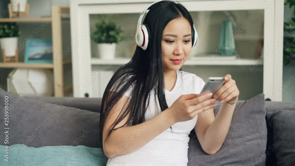 Asian young lady with beautiful long black hair is listening to music through headphones and using smartphone enjoying leisure time at home. Youth and technology concept. Asian young lady with beautiful long black hair is listening to music through headphones and using smartphone enjoying leisure time at home. Youth and technology concept.