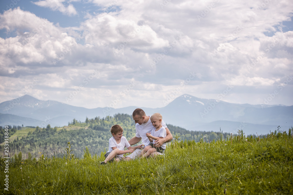 Happy father with his two young sons sitting on the grass on a background of green forest, mountains and sky with clouds. Friendship concept.