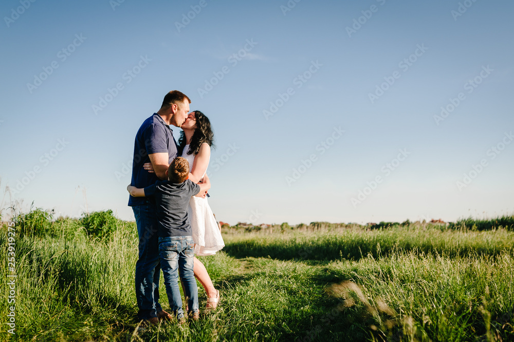 The son hugging parents who kissing on nature. Mom, dad and boy walk in the grass. Happy young family spending time together, outside, on vacation, outdoors. The concept of family holiday.