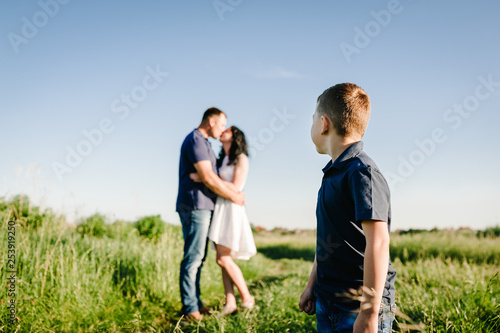 The son looks back at the parents who kiss in nature. Mom, dad and boy walk in the green grass. Happy young family spending time together, outside, on vacation, outdoors.