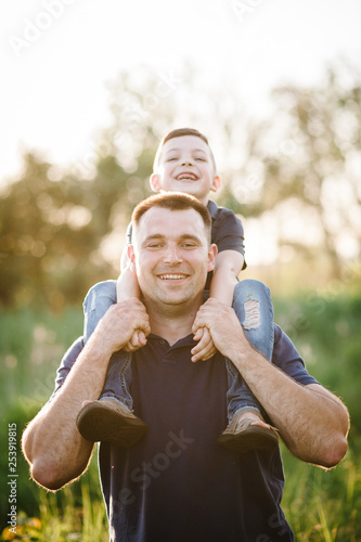 Portrait of happy father carrying on shoulders son on summer vacation. Father's Day. Dad and son playing in the park at the sunset time. People having fun on field. Concept of friendly family. 
