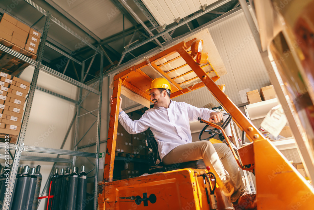 Smiling Caucasian worker in white uniform and protective helmet on head driving forklift in warehouse.