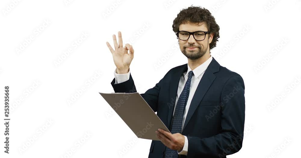 Handsome Caucasian businessman in the glasses, suit and tie looking at the documents in his hands and doing ok gesture to the camera on the white wall screen. Handsome Caucasian businessman in the glasses, suit and tie looking at the documents in his hands and doing ok gesture to the camera on the white wall screen.