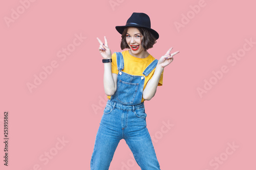 Portrait of excited beautiful young woman in yellow t-shirt and blue denim overalls with makeup and black hat standing and looking with victory or peace sign. studio shot isolated on pink background.