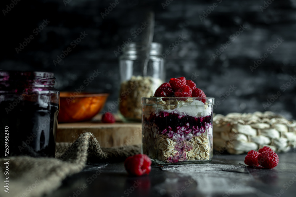 view on a dark, wooden table top, on which stands in a glass jar fresh yogurt with cereal, pits and raspberries, and ingredients for making it view on a dark, wooden table top, on which stands in a glass jar fresh yogurt with cereal, pits and raspberries, and ingredients for making it