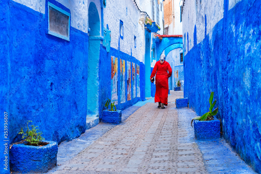 Unrecognizable Moroccan woman in red traditional clothes (jellaba) walking on a street in Medina of Chefchaouen, Morocco, small town in northwest Morocco known for its blue buildings