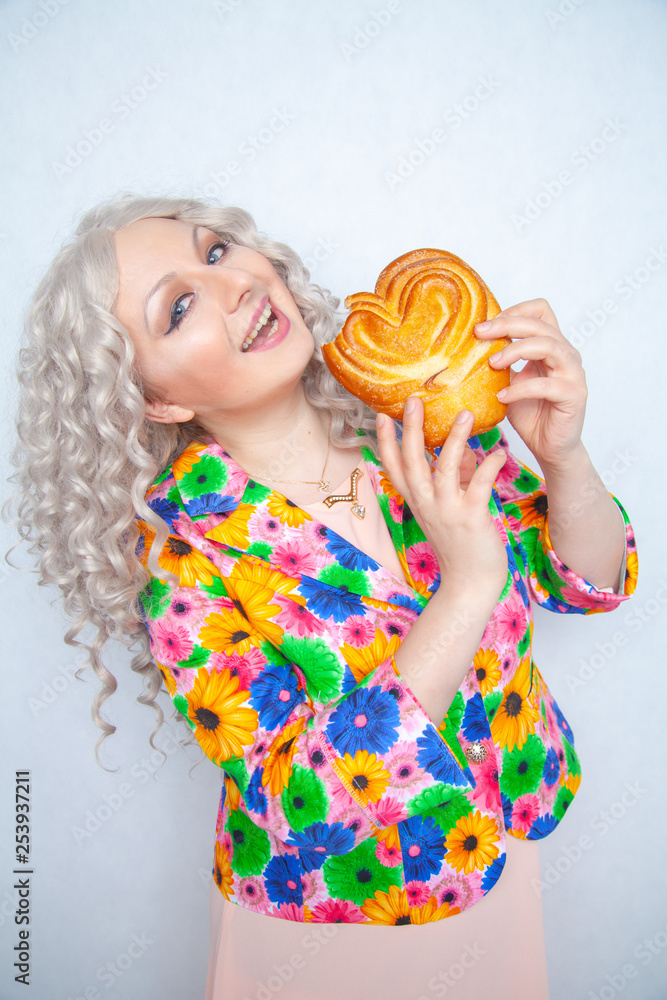 cute chubby girl with white curly hair in a summer jacket with flowers holds a sweet pastry bun in the shape of a heart