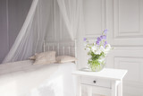 Glass vase with lilac and white floweers  in light cozy bedroom interior. White wall, bed with white linen, light blanket or plaid and pillows.