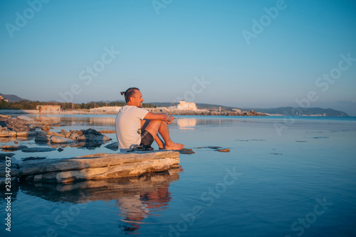 A young man sits on a stone by the sea.
