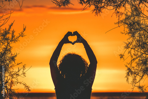 Silhouette image of a young woman making heart with hands on a beautiful summer evening Back view of a girl fingers like hearts sunset in background and branches that frame Enjoyable romance feelings