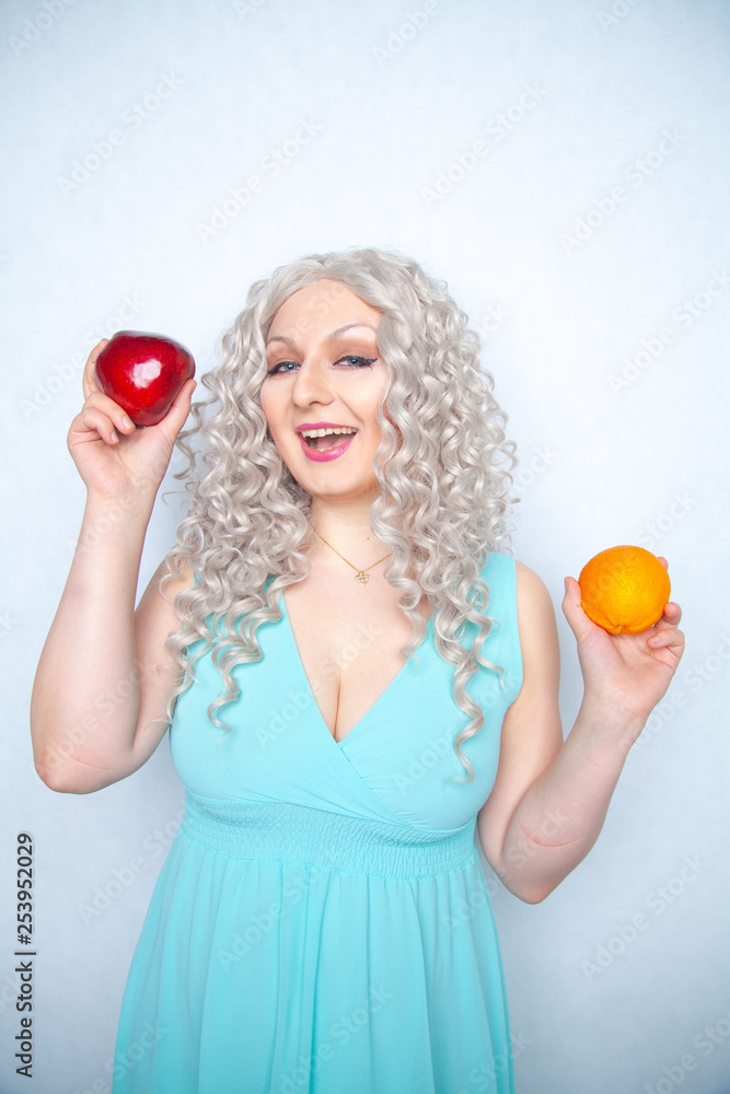 cute young girl with curly white hair and plump figure holding apple and orange fruit on studio background cute young girl with curly white hair and plump figure holding apple and orange fruit on studio background