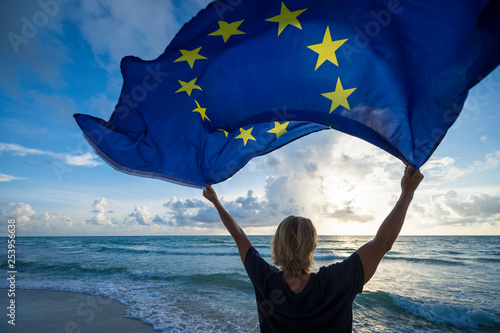 Fluttering European Union flag flying in bright morning sunlight held up by man with blond hair standing on empty Mediterranean beach