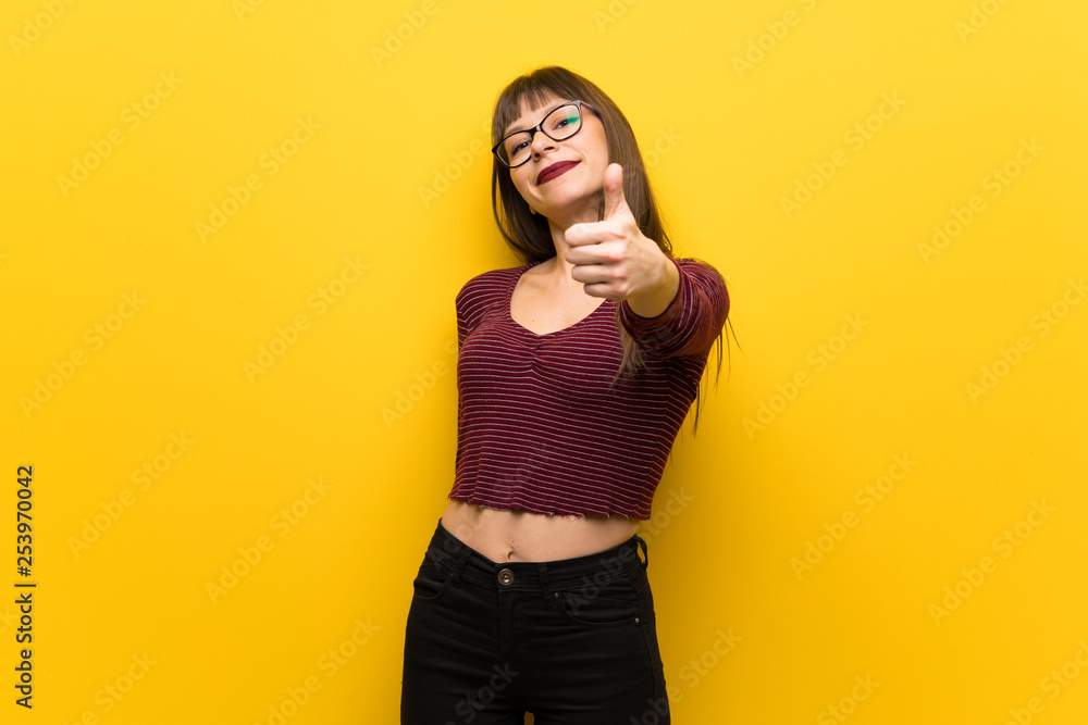Woman with glasses over yellow wall with thumbs up because something good has happened Woman with glasses over yellow wall with thumbs up because something good has happened