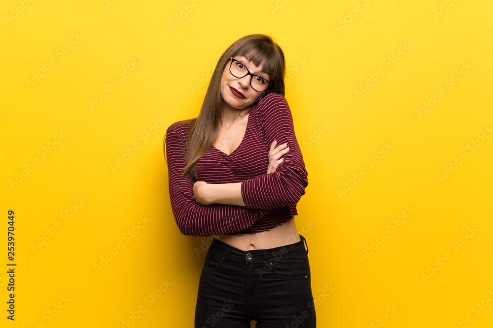 Woman with glasses over yellow wall making doubts gesture while lifting the shoulders Woman with glasses over yellow wall making doubts gesture while lifting the shoulders