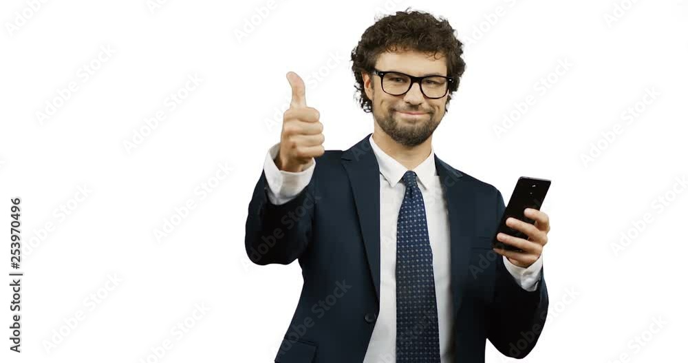 Caucasian smiled man in glasses, suit and tie giving his thumb up while scrolling and typing on the smartphone on the white screen background. Caucasian smiled man in glasses, suit and tie giving his thumb up while scrolling and typing on the smartphone on the white screen background.