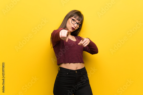 Woman with glasses over yellow wall points finger at you while smiling