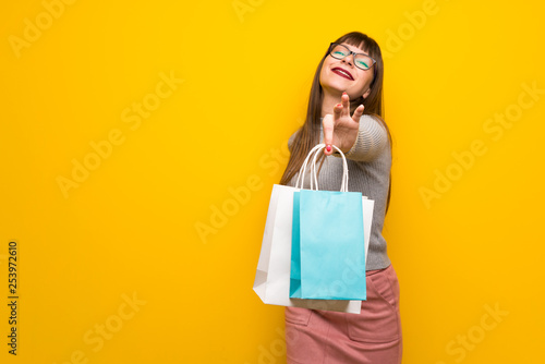 Woman with glasses over yellow wall holding a lot of shopping bags