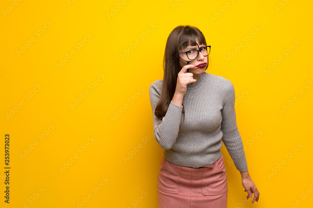 Woman with glasses over yellow wall taking a magnifying glass and looking through it Woman with glasses over yellow wall taking a magnifying glass and looking through it