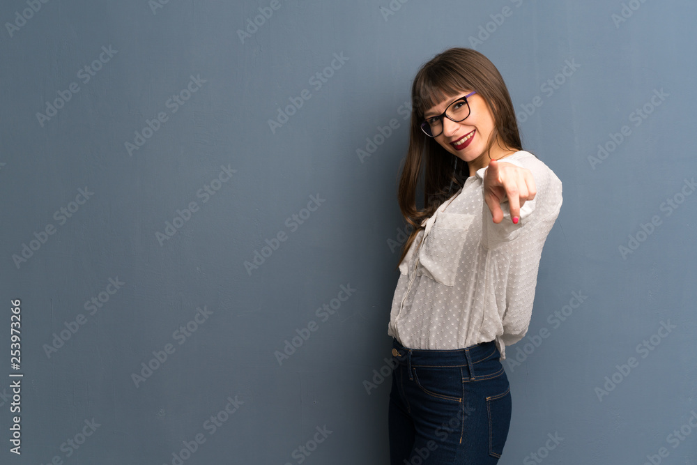 Woman with glasses over blue wall points finger at you with a confident expression Woman with glasses over blue wall points finger at you with a confident expression