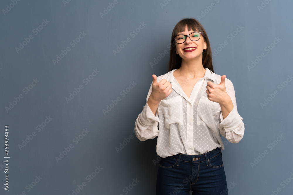 Woman with glasses over blue wall giving a thumbs up gesture with both hands and smiling Woman with glasses over blue wall giving a thumbs up gesture with both hands and smiling