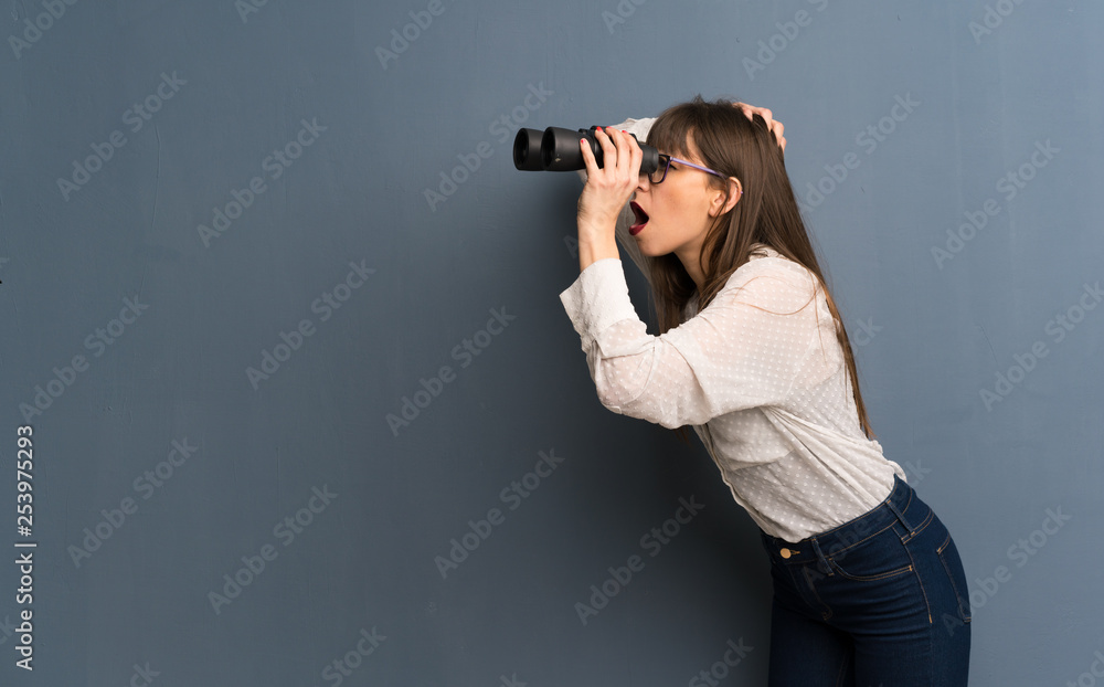 Woman with glasses over blue wall and looking in the distance with binoculars Woman with glasses over blue wall and looking in the distance with binoculars