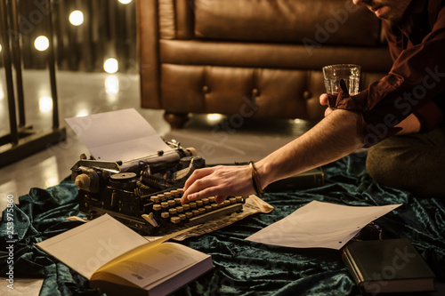 lateral view of man's hand typing on vintage typewriter on a piece of newspaper and dark blue velvet fabric on the floor in a dark room with light bulbs near a brown leather sofa