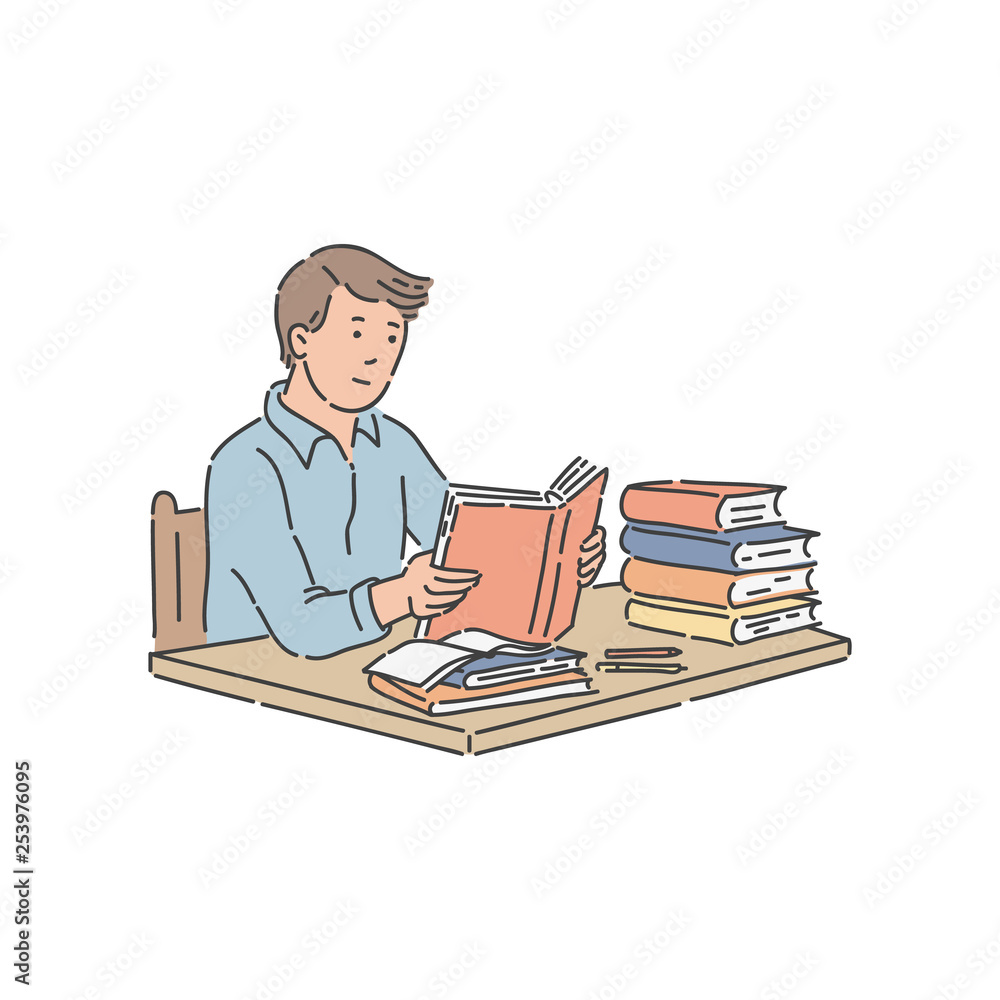 Male student sitting at table with pile of books and reading.
