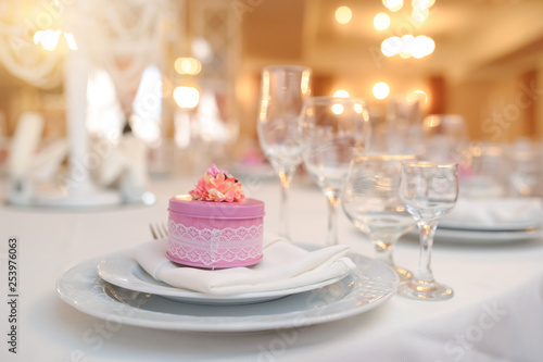 close up photo of knife and fork in a folded napkin on two plate near a set of glasses and a pink round gift box with a flower on the top on the napkin