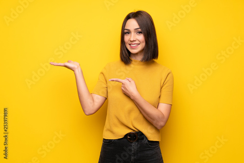 Young woman over yellow wall holding copyspace imaginary on the palm to insert an ad
