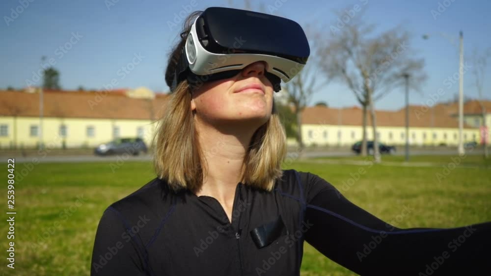 Happy sporty girl using virtual reality headset outdoor. Handheld shot of beautiful smiling young woman in sportswear using vr headset in park at sunny day. Technology concept Happy sporty girl using virtual reality headset outdoor. Handheld shot of beautiful smiling young woman in sportswear using vr headset in park at sunny day. Technology concept