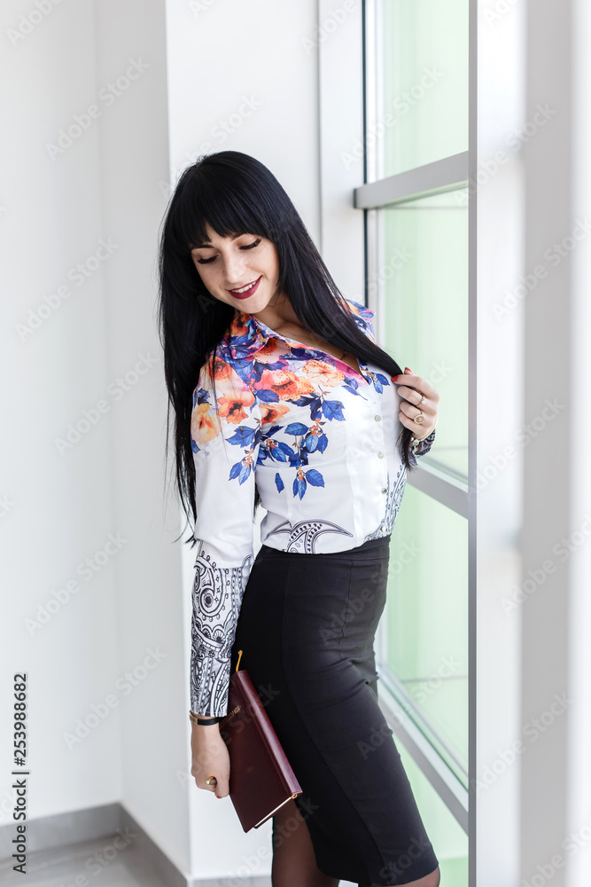 Young Attractive smiling brunette woman with long hair standing near the window in office, holding a notebook. Young Attractive smiling brunette woman with long hair standing near the window in office, holding a notebook.