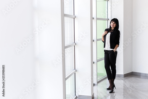 Young  happy brunette woman dressed in a black business suit standing near the window in a office  smiling  looking at camera.