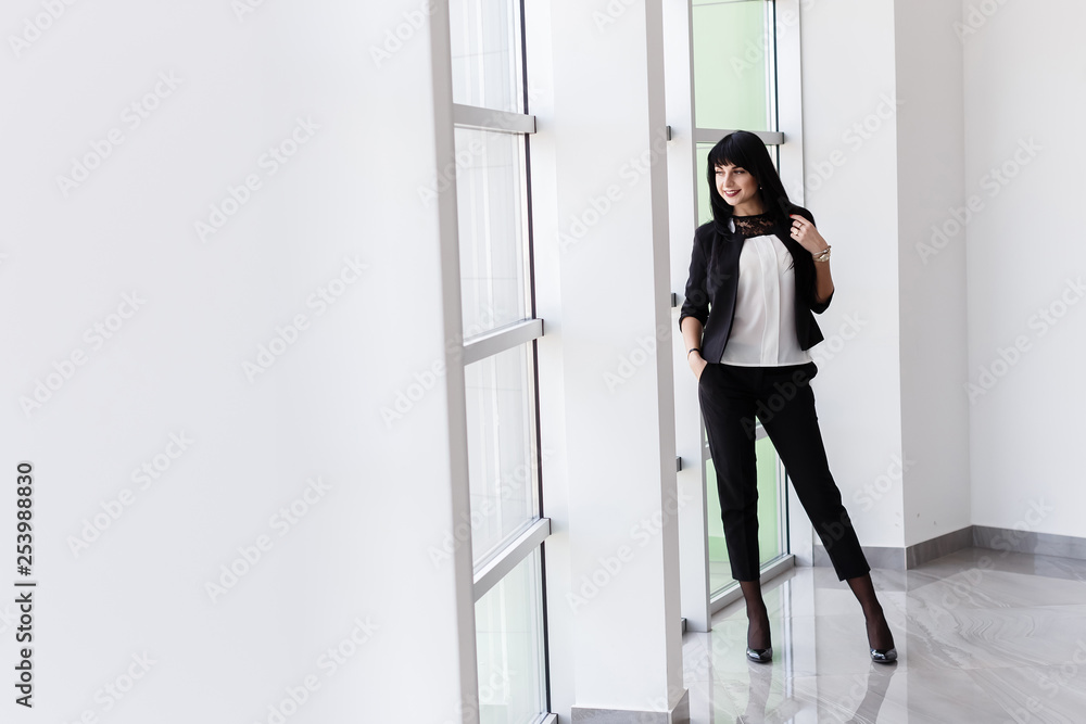 Young Attractive happy brunette woman with long hair standing near the window in office, smiling, looking out to window. Young Attractive happy brunette woman with long hair standing near the window in office, smiling, looking out to window.
