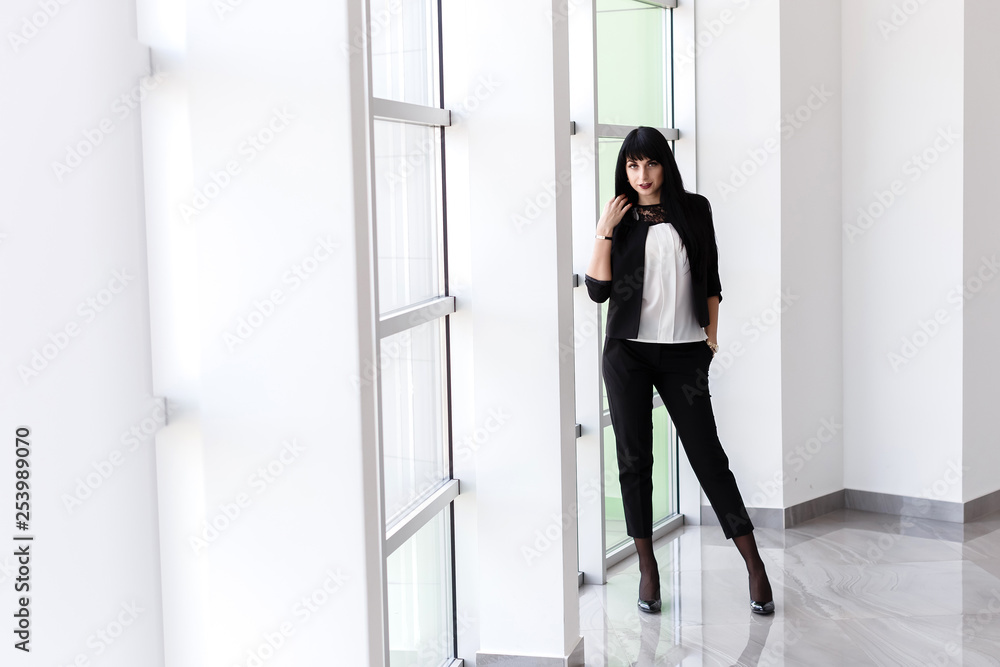 Young Attractive serious brunette woman dressed in a black business suit standing near the window in office, looking at camera. Young Attractive serious brunette woman dressed in a black business suit standing near the window in office, looking at camera.
