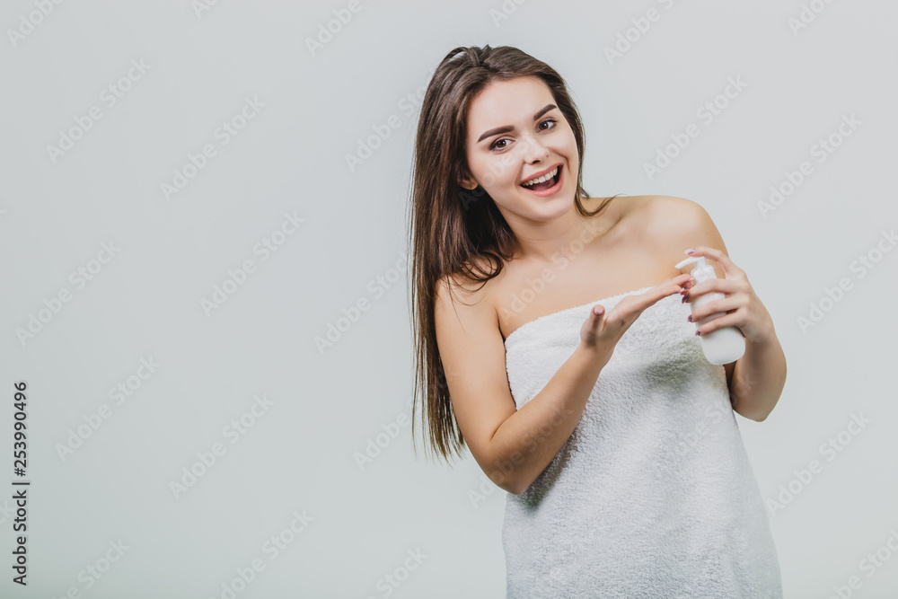 Fototapeta premium Model look is a beautiful young twin in a white terry towel. Applying a body lotion on her shoulder and smiling while standing on a white background.