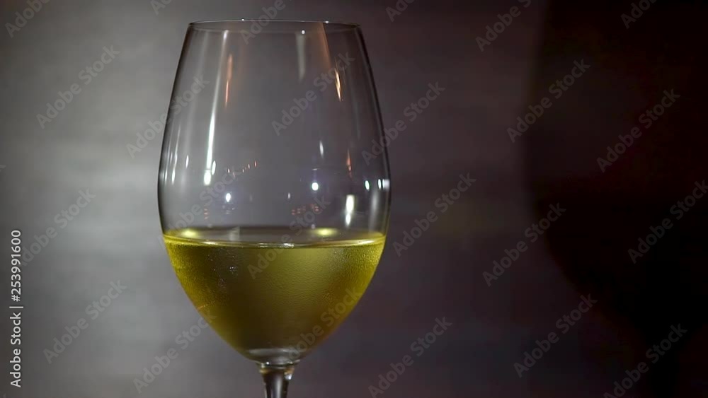 Camera rotates slowly, clockwise around single wine glass, half filled with white wine, against dark background