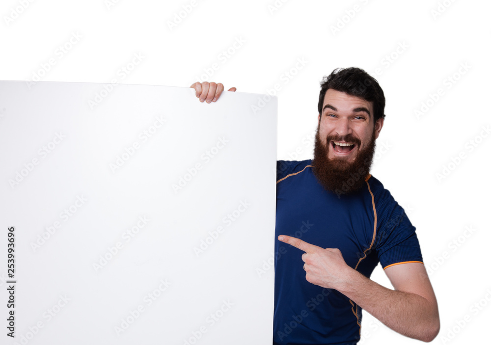 Young bearded man hiding behind white sheet and pointing on it on white wall background Young bearded man hiding behind white sheet and pointing on it on white wall background