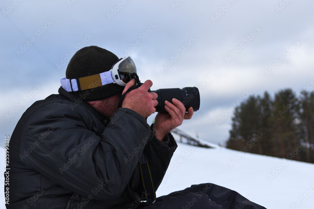 a photographer in ski goggles with a SLR camera and a long-focus lens takes pictures on the mountains of Vedashin