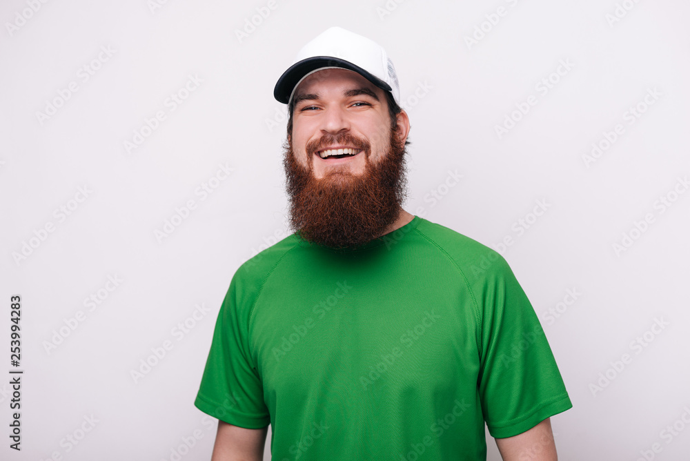 Portrait of bearded guy in green tshirt and hat, looking and smiling at camera standing over  white background