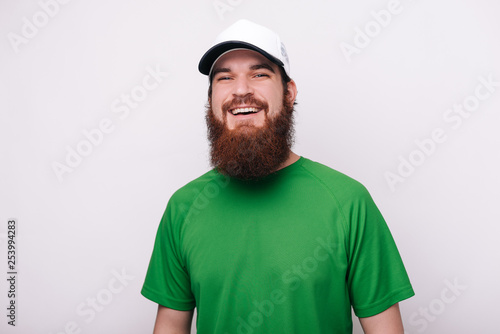 Portrait of bearded guy in green tshirt and hat, looking and smiling at camera standing over white background