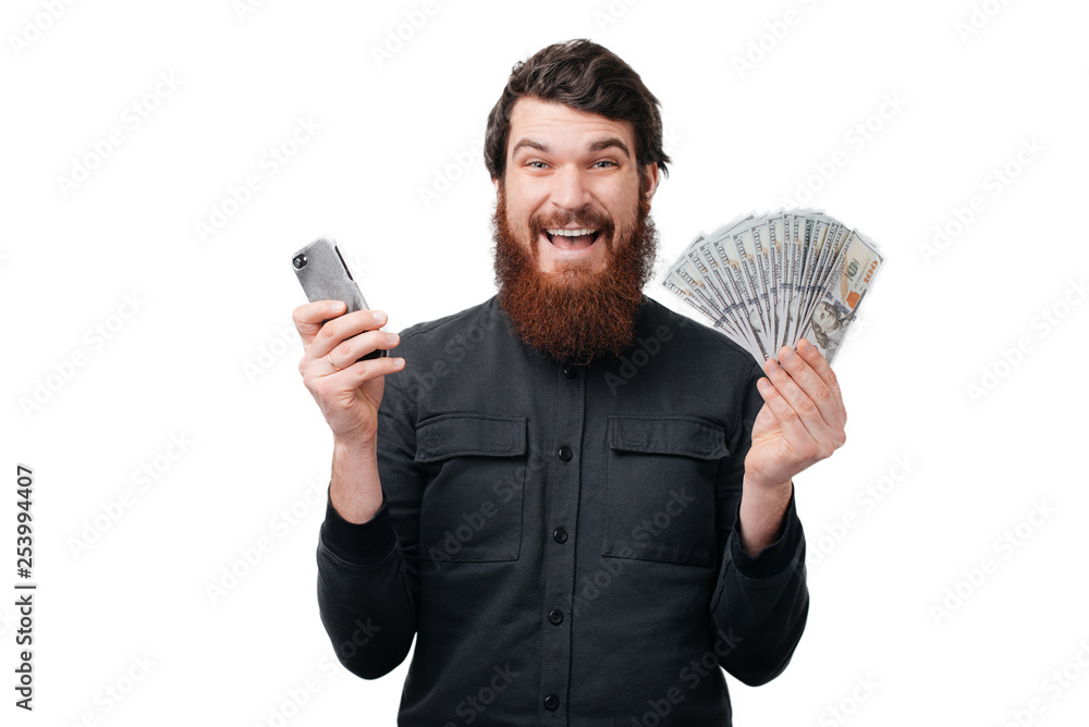 Excited bearded man in casual shirt holding lots of money in dollar currencys and cell phone in hands isolated over white wall Excited bearded man in casual shirt holding lots of money in dollar currencys and cell phone in hands isolated over white wall